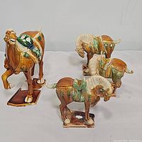 Four vintage ceramic Tang Dynasty style horses, including one large horse with raised leg and three smaller horses with bowed heads, all glazed in brown, green, and white.
