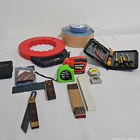 Photo showing wide array of hand tools including measuring tapes, foldable knives or saws, wire coil, matches, chalk sticks, brass fasteners, and other small hardware items