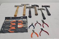 Full lot showing six hammers, pliers kit sealed with six orange handle pliers, and five loose pliers of varied types and colors.