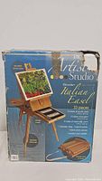 Front of box showing wooden Italian easel, paint tubes, brushes, canvas, palette, shoulder strap, and storage drawer.