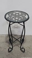 Full view of the metal plant stand showing its height, scroll legs, and round top with glass insert and ornate metal design.