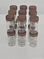 Photo showing 18 clear glass spice jars with wooden tops arranged in rows. Jars are labeled with various spices including five spice, ginger, and sage.