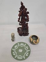 Photo showing all four items together on a white surface: Chinese wood carving statue, African bust sculpture, round glazed vase, and Wedgwood jasperware plate.