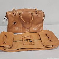 Two tan leather travel bags displayed, one carry-on upright and one duffel bag folded flat, showing handles and logo patches.