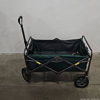 Side view of the collapsible utility cart showing metal frame, fabric body, two large front wheels, and retractable handle.
