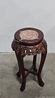 Full view of the ornate dark wood carved plant stand with round marble top. Stand has four legs with cross braces and detailed carved apron.