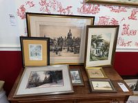 Photo showing a collection of framed etchings and watercolors arranged on a wooden surface and against a backdrop wall, depicting buildings, ducks, and landscapes.