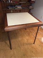 Card table showing full view with wood frame and light cushioned top in basement setting.