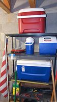 Three Igloo coolers stacked or arranged on black metal shelving, with the largest red and white cooler on top, middle shelf has blue and white thermos and a Playmate cooler, largest blue and white cooler on bottom shelf. US flag leaning nearby. Photo shows condition and arrangement of coolers, thermos, and flag.