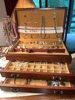 Jewelry box open showing multiple compartments and drawers filled with assorted costume jewelry including earrings, necklaces, bracelets, and pins.