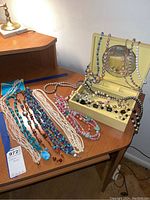 Image of assorted bead necklaces arranged on a wooden table with a yellow vintage jewelry box containing more necklaces, some draped over the box. The jewelry box has a mirror inside the lid.
