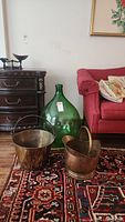 Green glass jug and two brass pails displayed on a patterned rug, with furniture in the background.