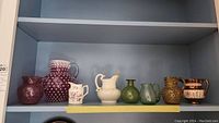 Shelf display of assorted creamers and pitchers including purple hobnail glass pitchers, green glass pitchers, and white ceramic items