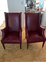 Front view of two matching mid-century modern wingback chairs with red upholstery and wooden legs on carpeted floor.