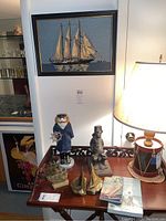 Photo showing the assembled nautical items on a table: framed needlepoint sailboat art on the wall, captain figurine with ship wheel, brass ship and sailboat bookends, brass ship figurine, nautical books, and a lamp shaped like a drum.