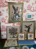 Framed needlepoint artworks displayed including bird scenes, a tree with berries, and sewing themed piece with text. Various frame sizes and styles visible.