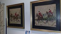 Two framed needlepoint fox hunt wall art pieces mounted on the wall, showing detailed scenes with riders in red jackets, horses, and hounds.