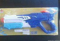 Blue, white, light blue water gun sealed in yellow-backed packaging with orange trigger and nozzle tip.