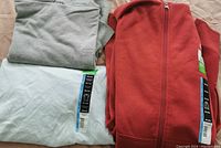 Three men's 3XL tops folded and stacked: light gray polo golf shirt, light blue T-shirt with tags, and red full zip-up hoodie with tags