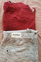 Two folded pairs of jogging pants, one red with black speckled pattern and one light gray with black speckled pattern showing the Jeaniebogie label on the gray pair.