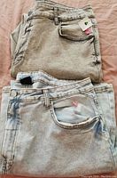 Top view of two pairs of folded light grey acid wash denim jeans showing front pockets and tags on one pair.