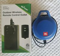 Photo of BN-LINK outdoor wireless remote control outlet in box and blue JBL mini speaker side by side on white textured surface.