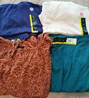 Four women's tops folded and displayed, three sleeveless shirts in navy blue, white, teal, and one rust-colored patterned blouse.