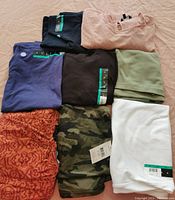 Eight women's 4XL tops folded on a flat surface. Includes plain, patterned, and camo tops with tags.