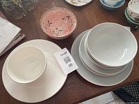 Photo showing stacked white dishes with concentric circle design, including bowls and plates, alongside plain white plates and the small pink floral bowl