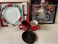 Grouped view of the lot showing boxed plate and utensil, two red mugs, glass Christmas bell shakers, small black ceramic plate, and glass etched platter in background