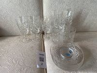 Photo showing two groups of crystal glassware on a beige patterned fabric background, consisting of goblets with stems and short tumblers stacked in two rows, plus a set of four small crystal plates arranged beside the glasses.