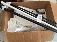 Open cardboard box containing folded sheer curtains, black curtain rods, and packaging for finials.