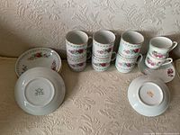 Lot of eleven espresso cups and nine saucers placed on a patterned cloth surface with visible floral patterns and manufacturer info on saucer bottom. Cups stacked and displayed.