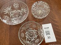 Three small pressed glass bowls, one with lid, all featuring decorative starburst and floral patterns on a wooden surface.