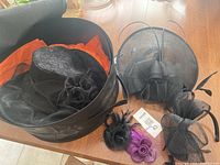 Large black hat with wide brim and fabric flower inside, alongside four fascinators including black and purple mesh flower designs with feathers