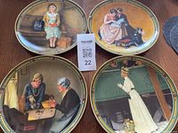 Four Norman Rockwell collector plates arranged on a wooden surface, showing various scenes including a woman sitting on a bench, a couple reading, and others.