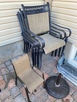 Four stacked adult outdoor chairs with black metal frames and beige mesh fabric. A single child outdoor chair with similar materials is positioned in front, alongside a black metal umbrella stand.