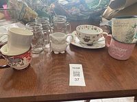 Image showing various mugs including clear glass mugs, owl-shaped creamer cup, and owl-themed ceramic mug on saucer alongside other mugs on wooden table.