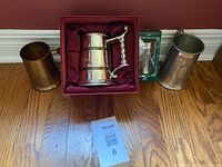 Four metal beer steins displayed on wood floor against red wall, including boxed silver stein, copper stein with etchings, and two plain brass/bronze steins