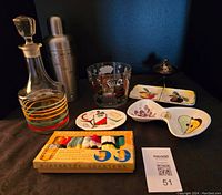 Overall view of the 1950s bar lot items arranged together on a dark surface, including decanter, cocktail shaker, ice bucket, coasters in box, dishes and lighter.