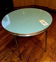 Full view of small round table with light blue atomic/starburst pattern laminate top, aluminum edge, and three metal legs.