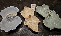 Photo of 3 leaf-shaped pottery serving dishes with handles on black surface; one pale blue with gold overspray, another pale blue-green with textured edges, and third beige with crackle glaze