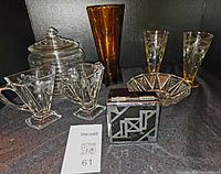 Assorted Art Deco glass and crystal items arranged on dark surface including cookie jar, amber vase, etched glasses, crystal bowl, and a decorative lidded box.