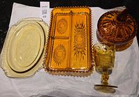 Set of four glassware items on black surface: large cream depression oval platter, amber divided dish, amber covered nut dish, yellow-gold goblet.