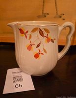 Front and side photos of a white Hall China pitcher with an orange and brown floral pattern, ribbed body texture, and gold accents on rim and handle taken indoors on a wooden surface.