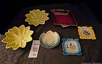 All six shallow dishes displayed on tablecloth: two large yellow leaf-shaped Carlton Ware dishes, green and white leaf dish, maroon and gold rectangular dish, two Japanese porcelain square dishes with painted designs.