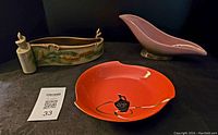 Photo of the lot showing the three items on a dark surface, including red lacquer dish, pink Hull swoop dish, and rectangular green glazed Beauceware planter with pinecone relief.