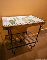 Full view of mid century serving cart with metal frame, lower shelf, casters and decorated glass top with botanical prints.