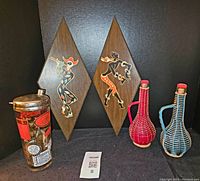Overall view of midcentury bar set showing cocktail shaker, two decanters with woven covers, and pair of dancing figure plaques.