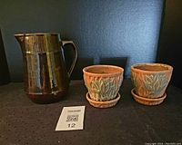 Overall view of the brown glossy pottery jug and two matte glazed jardiniere pots with matching saucers against a black background.